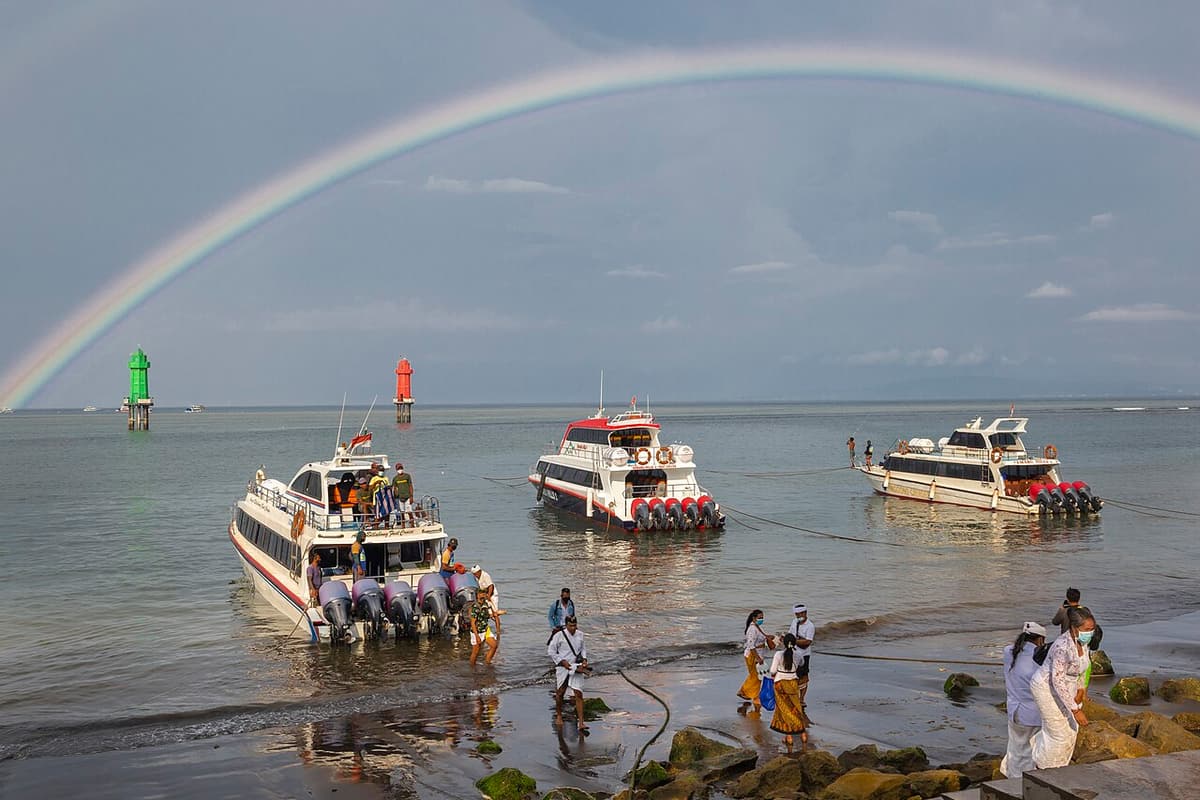 Des passagers rejoignent à pied, dans l’eau, les bateaux rapides à Sanur sous un arc-en-ciel